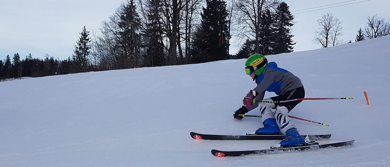 A skier is descending a snowy slope with skis lying on the ground in front. The background shows a forest with leafless trees.