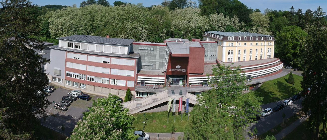 Eine Luftaufnahme eines Gebäudekomplexes, möglicherweise ein Hotel, mit einem Parkplatz und Flaggen davor. Bäume und eine Straße sind im Vordergrund.