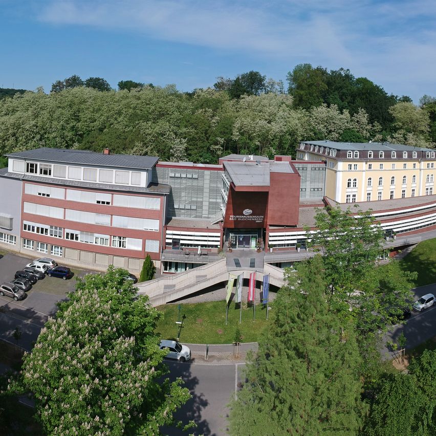 Eine Luftaufnahme eines Gebäudekomplexes, möglicherweise ein Hotel, mit einem Parkplatz und Flaggen davor. Bäume und eine Straße sind im Vordergrund.