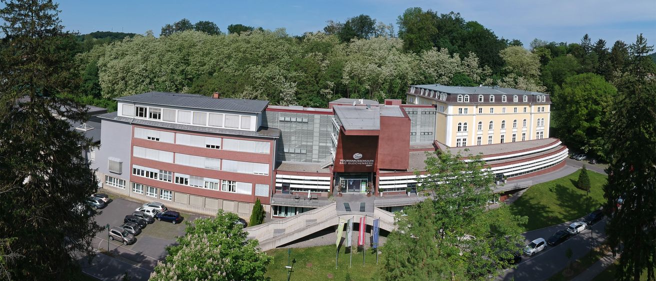 Eine Luftaufnahme eines Gebäudekomplexes, möglicherweise ein Hotel, mit einem Parkplatz und Flaggen davor. Bäume und eine Straße sind im Vordergrund.