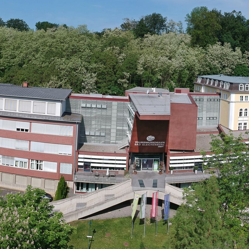 Eine Luftaufnahme eines Gebäudekomplexes, möglicherweise ein Hotel, mit einem Parkplatz und Flaggen davor. Bäume und eine Straße sind im Vordergrund.