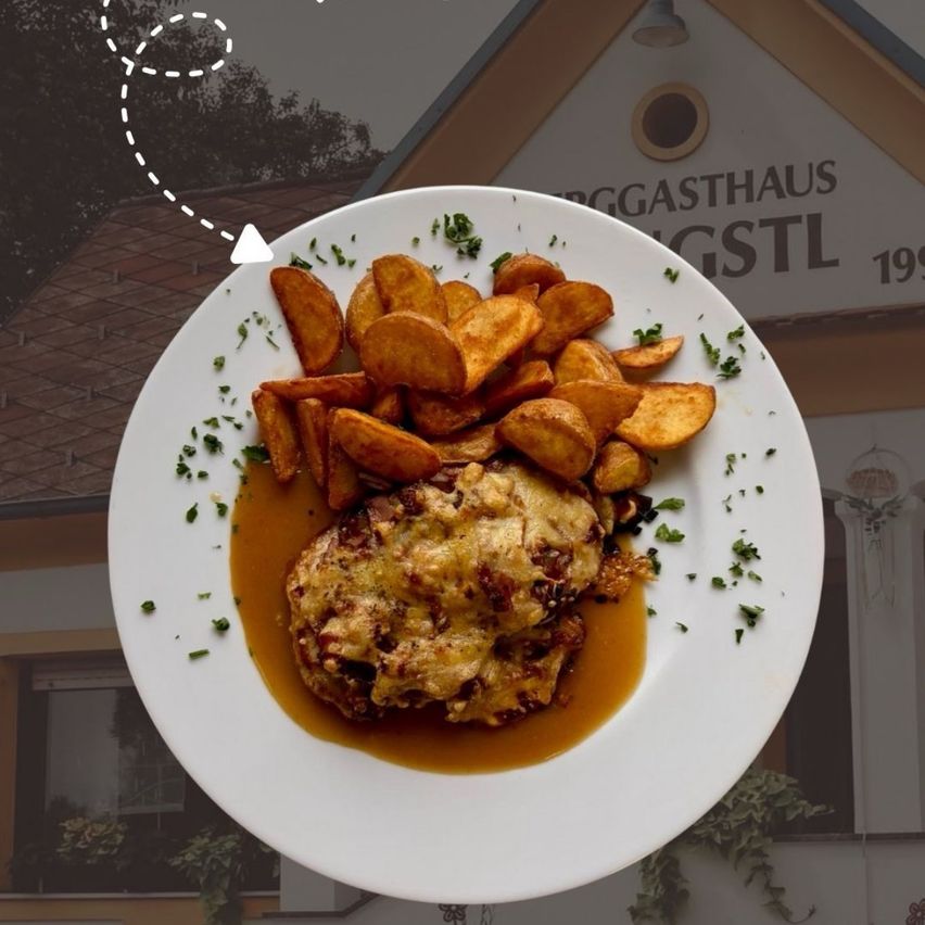 Ein weißer Teller mit einem Gericht mit goldener Kruste und einer Soße, begleitet von einer Menge Kartoffelspalten und Garnierungen. Der Teller steht auf einem Tisch vor einem Gebäude mit der Aufschrift 'EGGASTHAUS GSTL 19'.