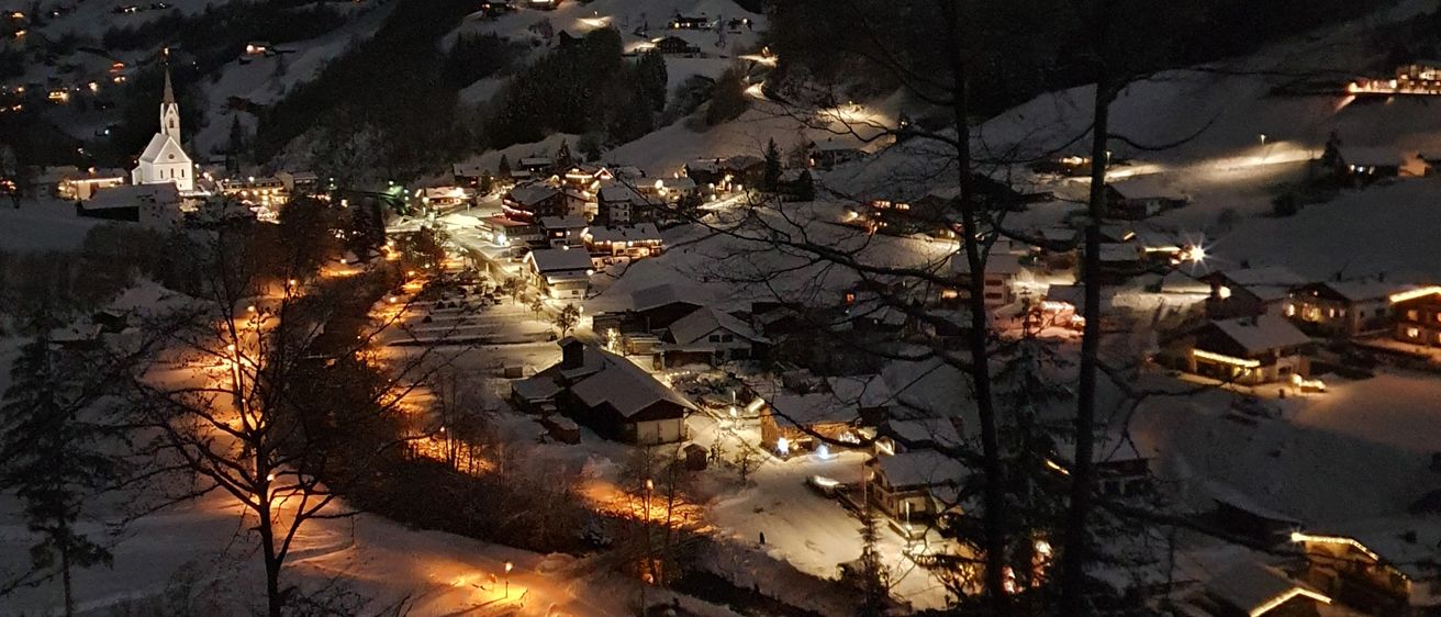 Eine Luftaufnahme eines verschneiten Bergdorfs bei Nacht, mit beleuchteten Häusern und einem Kirchturm sichtbar.