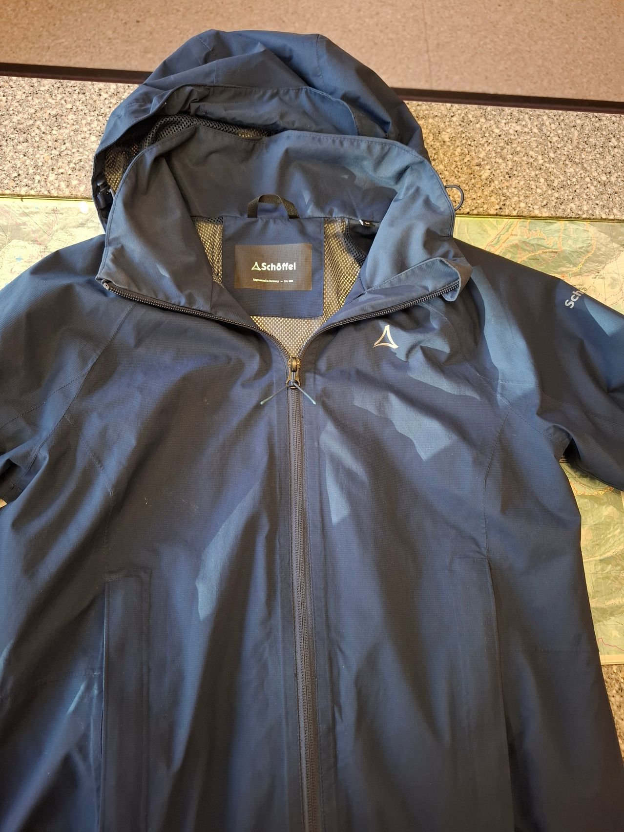 Eine wasserdichte navyblaue Jacke mit Kapuze und Reißverschluss. Die Jacke hat ein Netzpanel unter dem Reißverschluss und ein Markenetikett an der Kapuze.