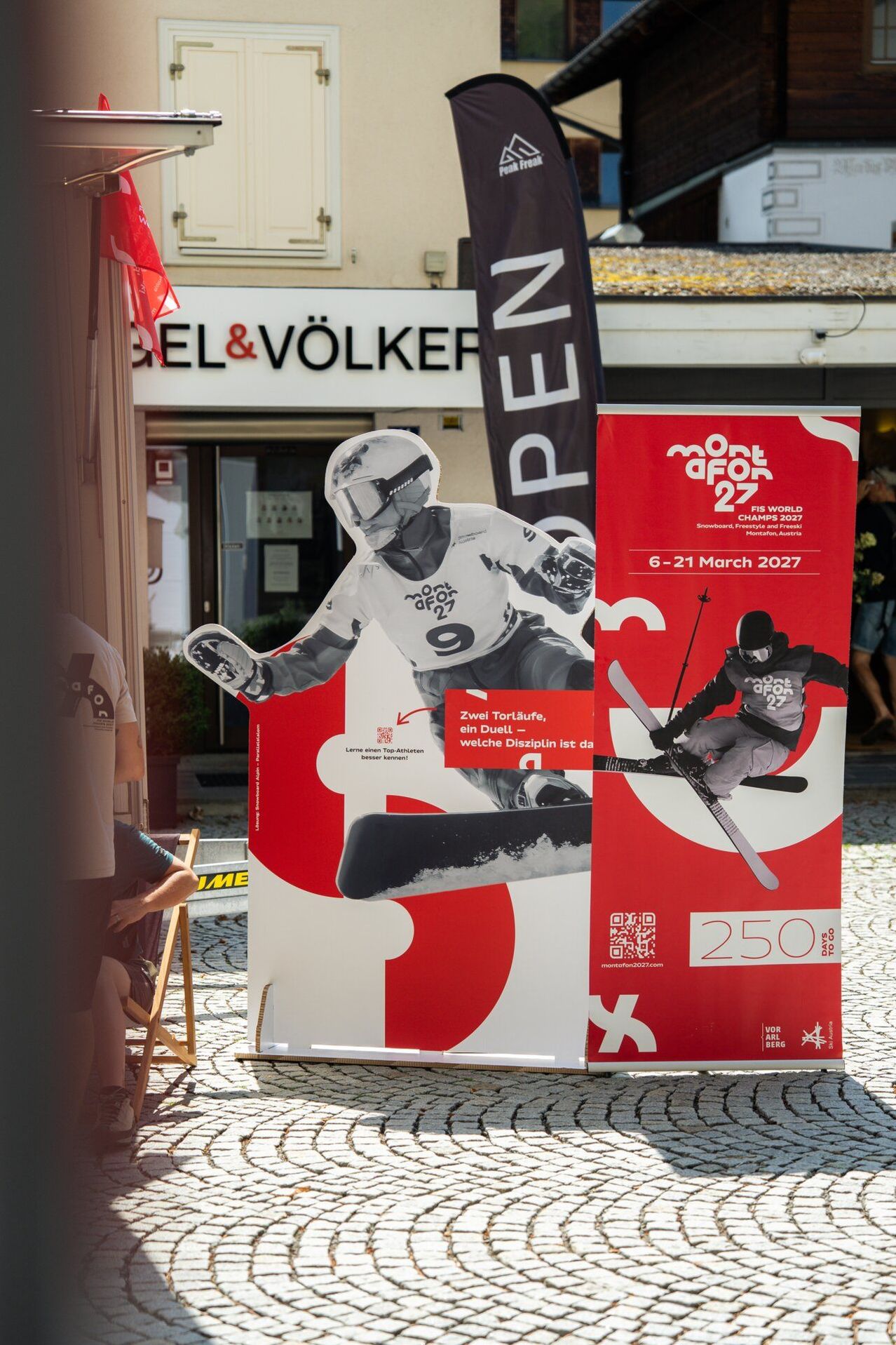 Ein großes Plakat für die FIS-Weltmeisterschaften im Snowboarding und Freestyle 2027 in Montafon, Österreich, mit einem Skifahrer und Snowboarder und dem Slogan 'Zwei Torläufe, ein Duell'. Das Plakat ist vor einem Gebäude mit einer roten Fahne angebracht.