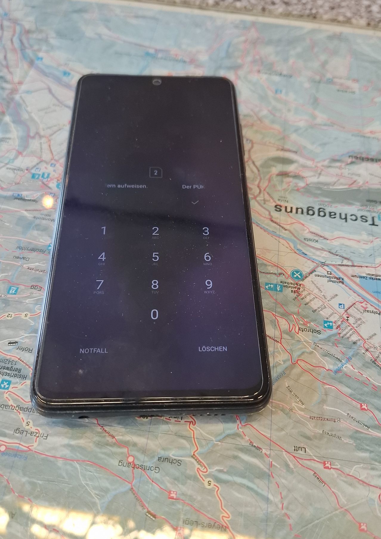 Ein Smartphone ist auf einer Karte geöffnet. Der Bildschirm des Telefons zeigt eine PIN-Eingabe mit den Ziffern 1 bis 9 und 0. Unter der PIN-Eingabe befinden sich Tasten für 'NOTFALL' und 'LÖSCHEN'. Die Karte hat verschiedene Bezeichnungen.