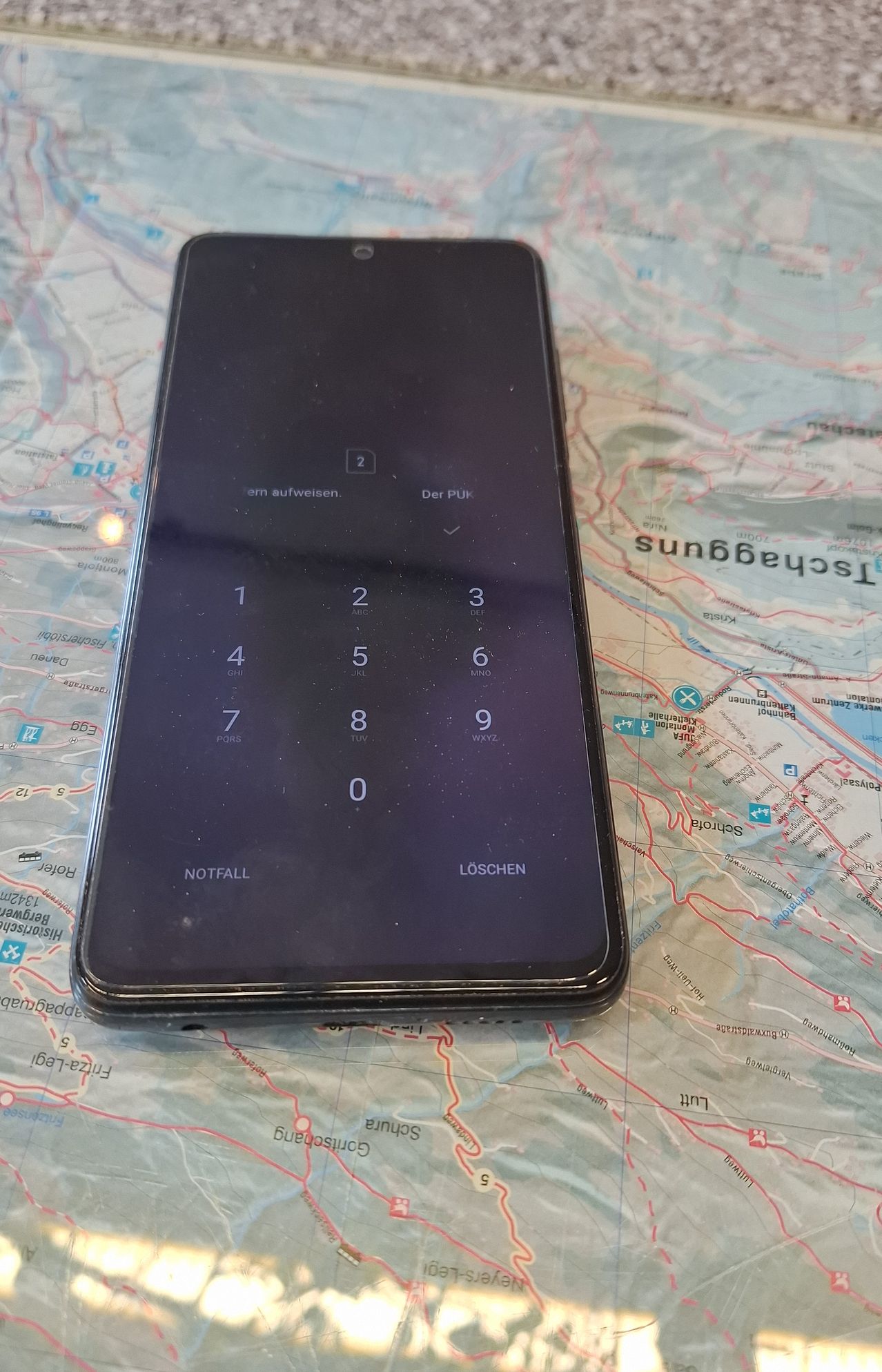 Ein Smartphone ist auf einer Karte geöffnet. Der Bildschirm des Telefons zeigt eine PIN-Eingabe mit den Ziffern 1 bis 9 und 0. Unter der PIN-Eingabe befinden sich Tasten für 'NOTFALL' und 'LÖSCHEN'. Die Karte hat verschiedene Bezeichnungen.