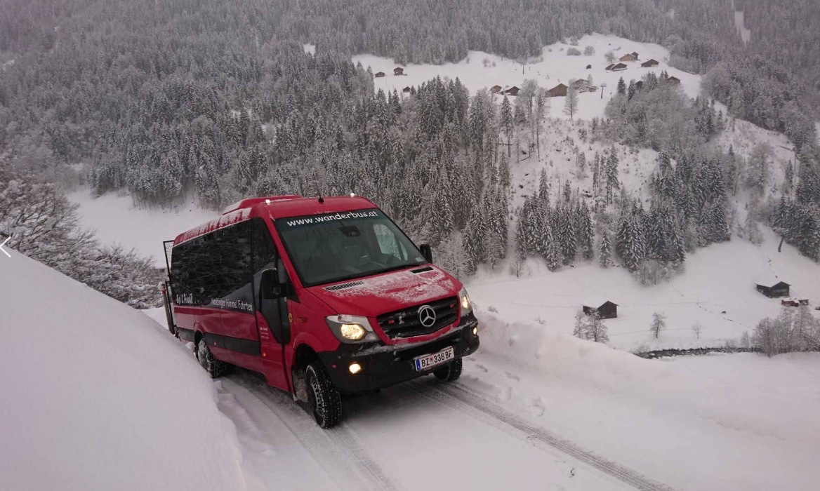 Ein roter Mercedes Sprinter fährt auf einer verschneiten Bergstraße, mit einer Website-Adresse auf der Windschutzscheibe und einem Nummernschild, das BZ 336 GF liest.