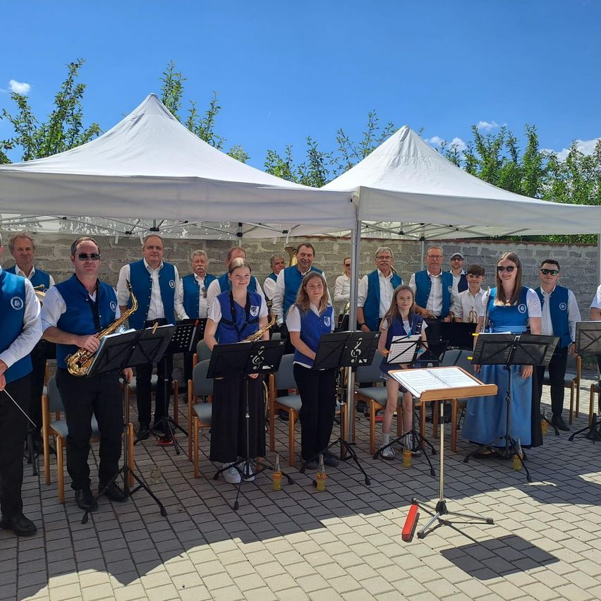 A group of musicians in blue vests and black pants stand under a white tent, holding instruments and music stands. They are likely preparing for a performance.