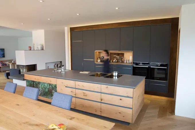 A modern kitchen with a large island, dark cabinets, and a fireplace in the background. There is a person standing inside the kitchen.