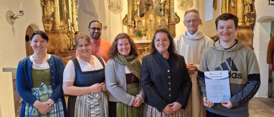Eine Gruppe von Personen, einschließlich eines Priesters, steht vor einem Altar und lächelt für ein Foto. Zwei Frauen halten Urkunden in den Händen, und ein Mann rechts trägt eine Adidas-Hoodie.