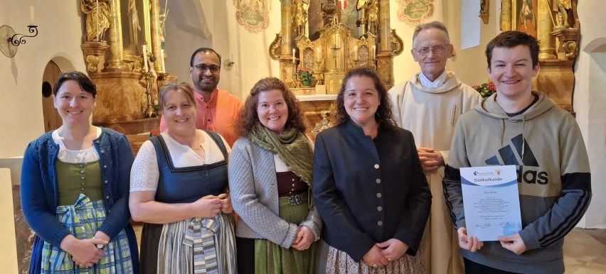 Eine Gruppe von Personen, einschließlich eines Priesters, steht vor einem Altar und lächelt für ein Foto. Zwei Frauen halten Urkunden in den Händen, und ein Mann rechts trägt eine Adidas-Hoodie.