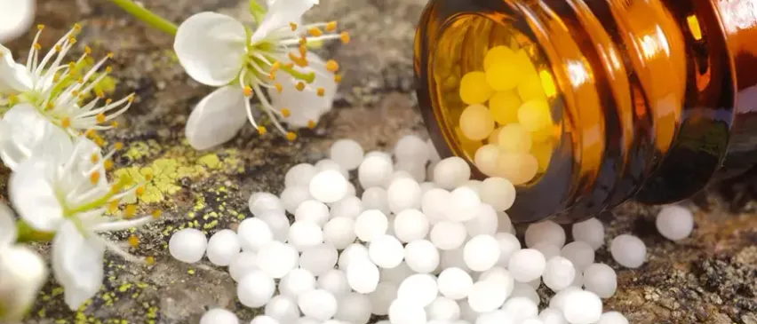 Eine braune Glasflasche mit einigen gelben und weißen Pillen ist geöffnet und verschüttet ihren Inhalt auf einer rauen Oberfläche. Im Hintergrund befinden sich weiße Blumen mit gelben Zentren.