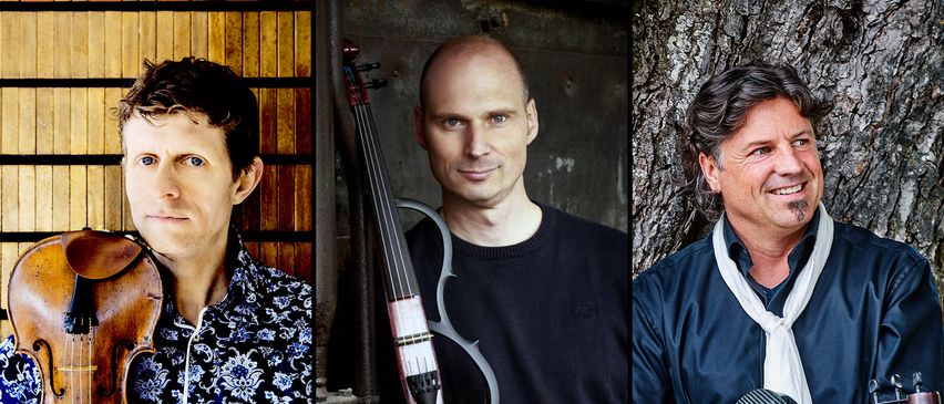 Ein Mann in einem schwarzen Pullover, der eine einzigartige Geige mit einem metallischen Bogen hält, steht vor einer Holzwand.