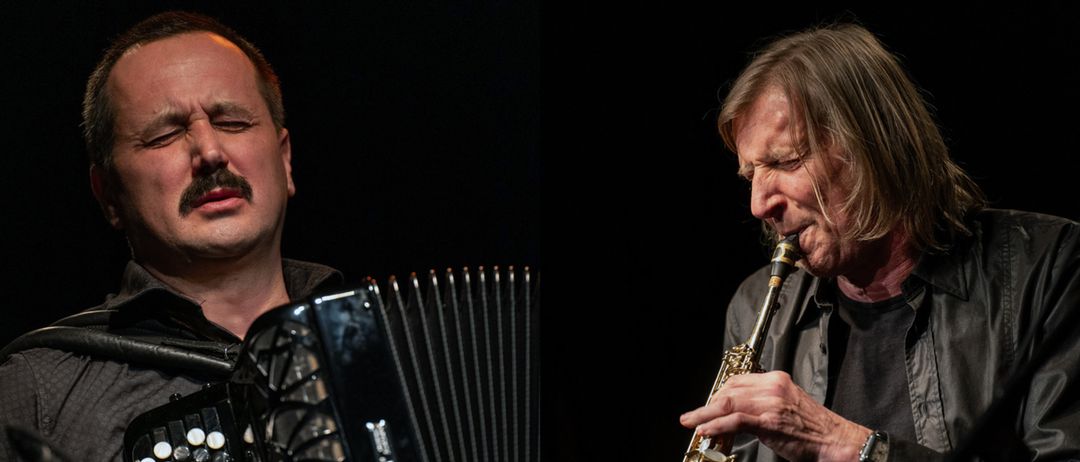 Ein Musiker spielt ein Saxophon, während ein schwarzes Akkordeon im Vordergrund steht. Der Musiker scheint sich intensiv auf seine Darbietung zu konzentrieren.