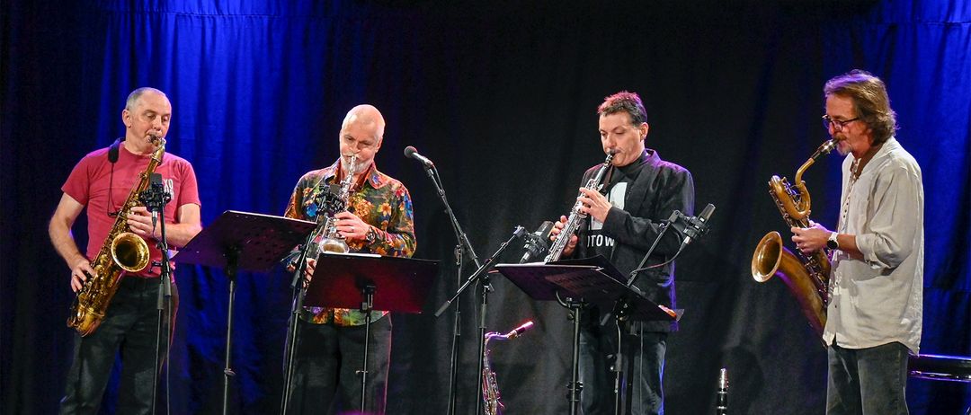 Zwei Musiker spielen auf der Bühne, einer mit einer Saxophon und der andere mit einer Klarinette. Beide tragen formelle Kleidung. Hinter ihnen steht ein Mikrofon auf einem Ständer für ihre Aufführung.