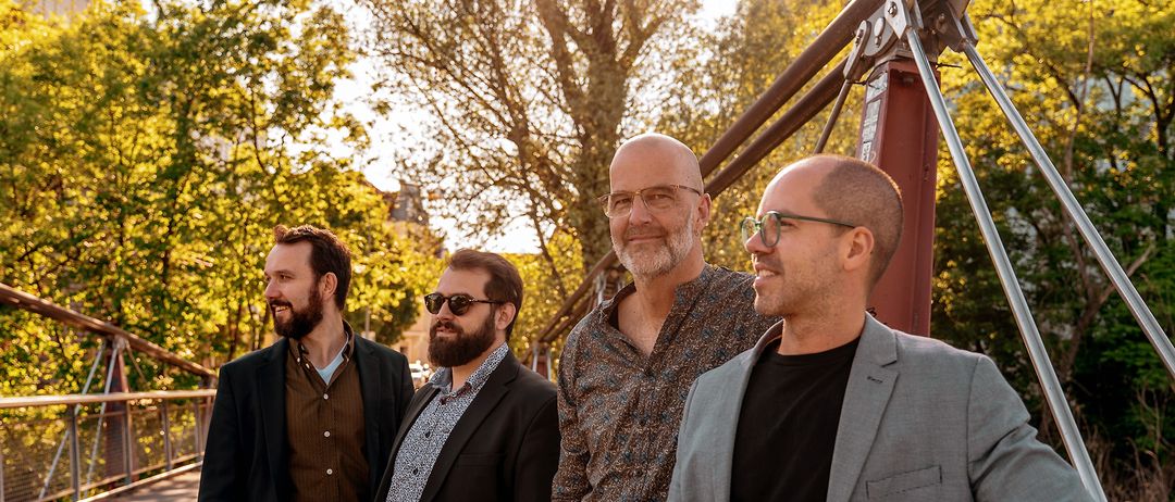 Vier Männer, schick gekleidet, stehen unter einem Baum in einem Park. Sie scheinen zu lächeln und nach vorne zu schauen. Der Hintergrund ist unscharf.
