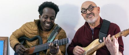 Zwei Männer spielen Gitarre, einer lächelt, während der andere eine Brille und einen Bart trägt. Sie scheinen sich in einer ungezwungenen, inländischen Umgebung zu befinden.