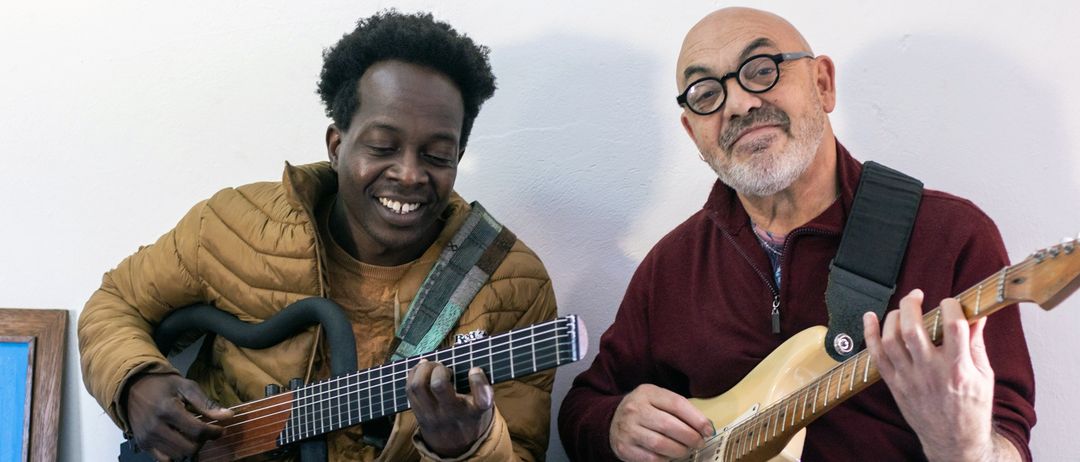 Zwei Männer spielen Gitarre, einer lächelt, während der andere eine Brille und einen Bart trägt. Sie scheinen sich in einer ungezwungenen, inländischen Umgebung zu befinden.