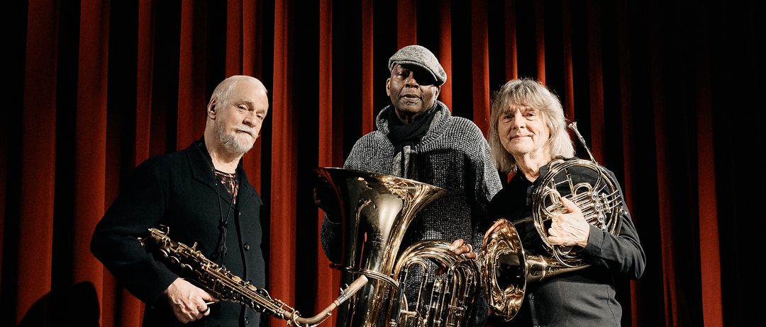 Drei Musiker stehen auf der Bühne vor roten Vorhängen, einer hält eine Saxophon und zwei halten Tubas. Der Mann links trägt einen schwarzen Mantel, der mittlere Mann trägt einen grauen Strickmütze und einen Schal.