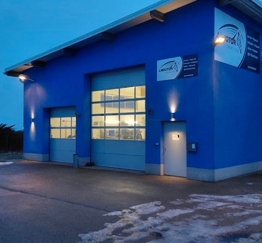 A blue building with a large garage door and a sign reading 'Lamotor'. The building has a modern design with glass windows and lights on the sides.