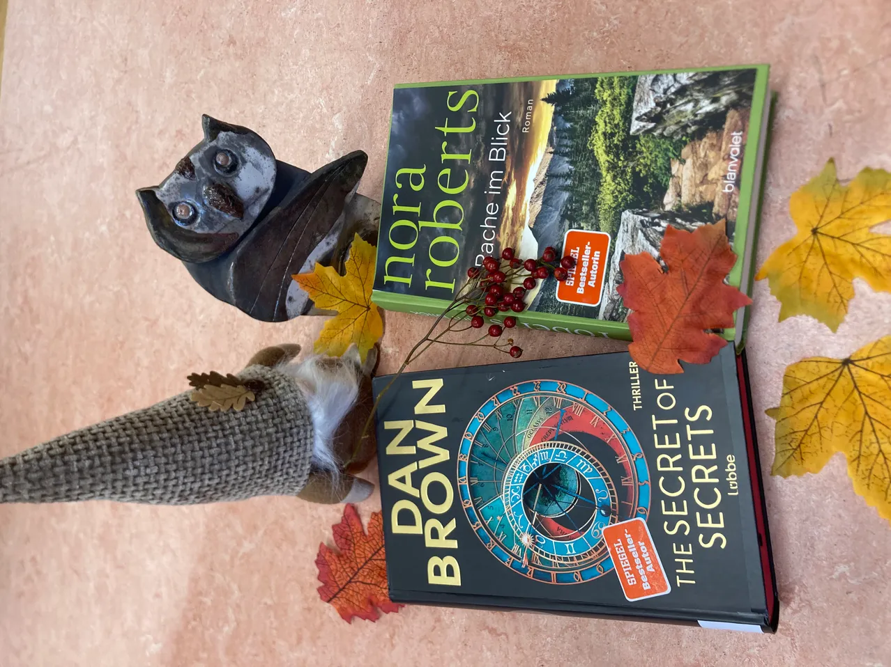 Zwei Bücher und eine fledermausartige Figur auf einer beigefarbenen Oberfläche, Herbstblätter herum, ein Buch von Nora Roberts und ein anderes von Dan Brown.