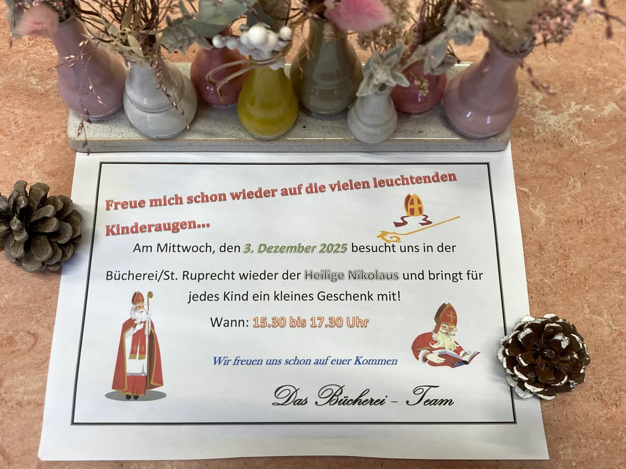 Ein Schild auf einem Tisch, umgeben von Vasen, lädt Kinder ein, die Bibliothek am 3. Dezember 2025 zu besuchen, wo der Heilige Nikolaus Geschenke bringt. Das Schild ist mit einer Zeichnung des Heiligen Nikolaus und einer Tannenzapfen verziert.