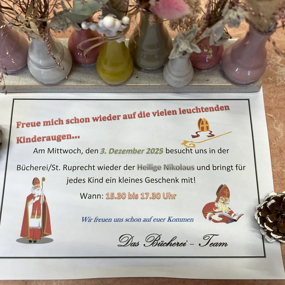 Ein Schild auf einem Tisch, umgeben von Vasen, lädt Kinder ein, die Bibliothek am 3. Dezember 2025 zu besuchen, wo der Heilige Nikolaus Geschenke bringt. Das Schild ist mit einer Zeichnung des Heiligen Nikolaus und einer Tannenzapfen verziert.