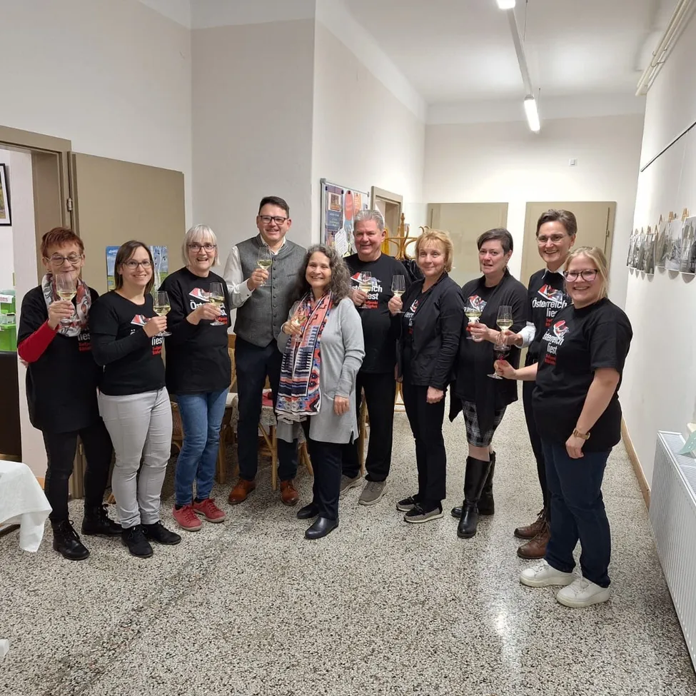 Eine Gruppe von Menschen steht zusammen in einem Raum, hält Weingläser in der Hand und lächelt für ein Foto. Sie tragen alle schwarze T-Shirts mit weißen Buchstaben.