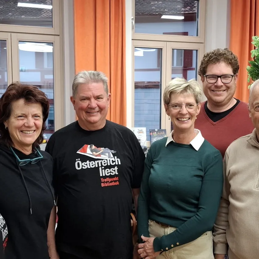 Fünf Menschen lächeln für ein Foto. Ein Mann in einem schwarzen Shirt mit dem Text 'Österreich liest' steht in der Mitte. Die anderen stehen auf beiden Seiten von ihm. Hinter ihnen sind zwei Fenster mit Vorhängen.