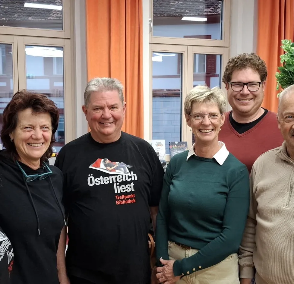 Fünf Menschen lächeln für ein Foto. Ein Mann in einem schwarzen Shirt mit dem Text 'Österreich liest' steht in der Mitte. Die anderen stehen auf beiden Seiten von ihm. Hinter ihnen sind zwei Fenster mit Vorhängen.