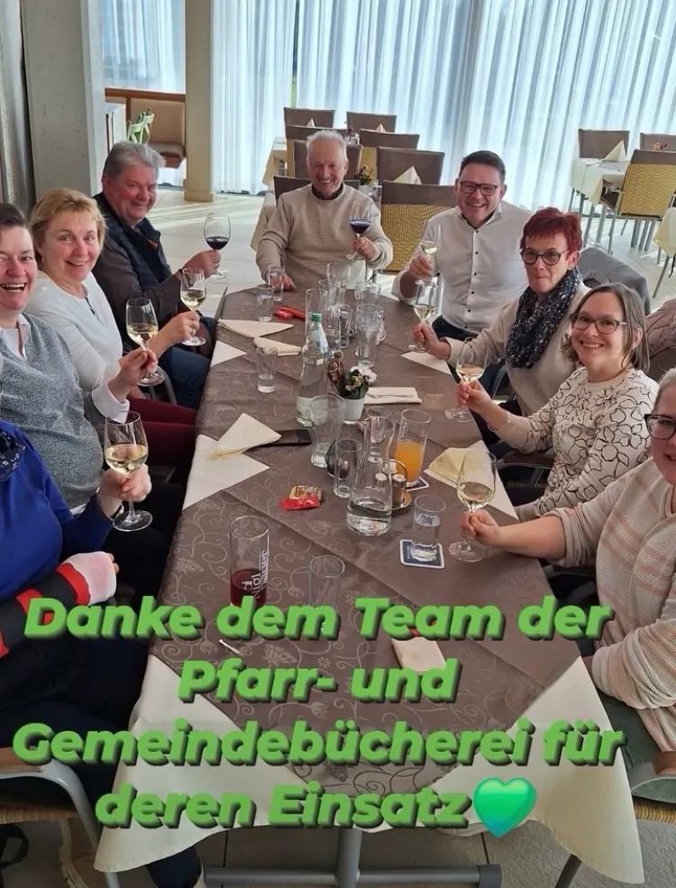 Eine Gruppe von Menschen sitzt um einen Tisch herum, lächelt und hält Weinglas in der Hand. Der Tisch ist mit Weingläsern, einer Flasche Wein und einer Vase mit Blumen gedeckt. Sie feiern wahrscheinlich ein Ereignis. Das Bild hat einen Wasserzeichen mit dem Text 'Danke dem Team der Pfarr- und Gemeindebibliothekerei für'.