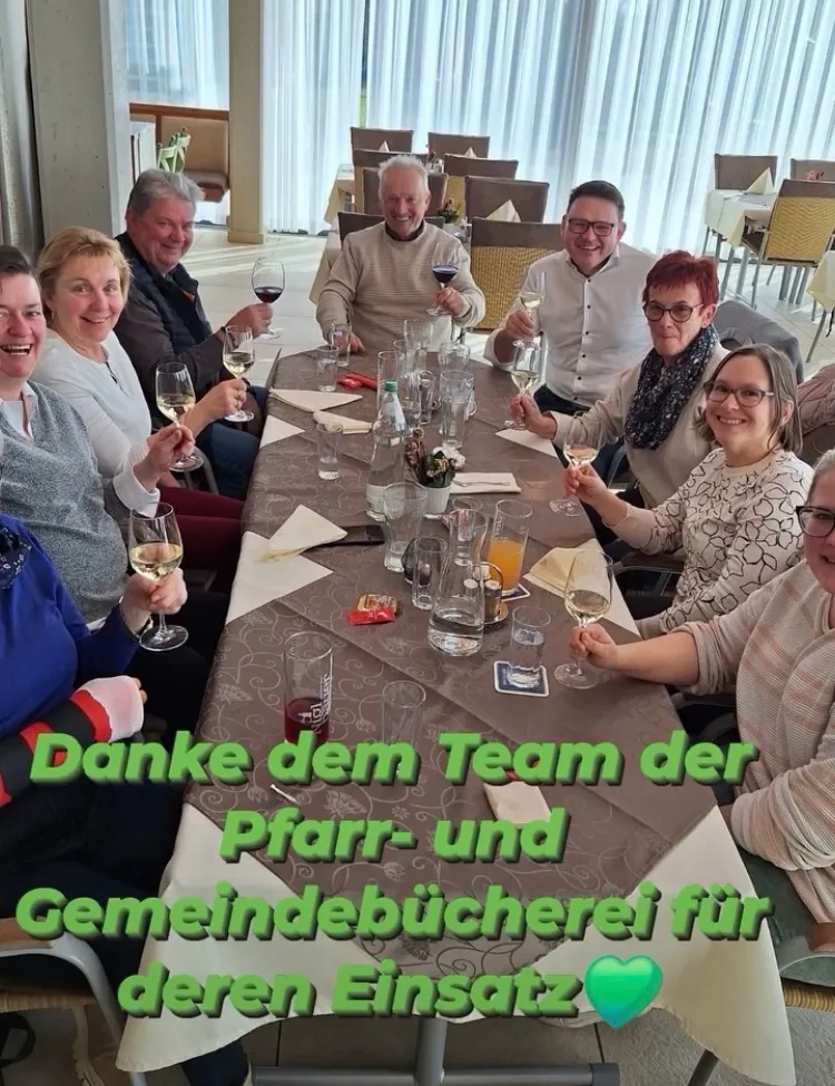 Eine Gruppe von Menschen sitzt um einen Tisch herum, lächelt und hält Weinglas in der Hand. Der Tisch ist mit Weingläsern, einer Flasche Wein und einer Vase mit Blumen gedeckt. Sie feiern wahrscheinlich ein Ereignis. Das Bild hat einen Wasserzeichen mit dem Text 'Danke dem Team der Pfarr- und Gemeindebibliothekerei für'.