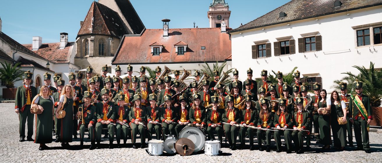 Ein Militärorchester in grünen Uniformen und Mützen posiert für ein Foto vor einem historischen Gebäude mit einem Uhrturm.