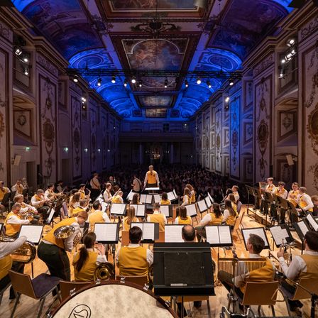 Ein Orchester in gelben Westen spielt auf der Bühne unter blauer Beleuchtung in einem großen Saal. Ein Dirigent steht davor, und das Publikum füllt die Plätze. Die Wände sind mit aufwendigen Verzierungen geschmückt.