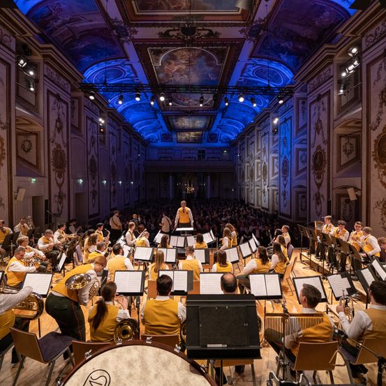 Ein Orchester in gelben Westen spielt auf der Bühne unter blauer Beleuchtung in einem großen Saal. Ein Dirigent steht davor, und das Publikum füllt die Plätze. Die Wände sind mit aufwendigen Verzierungen geschmückt.