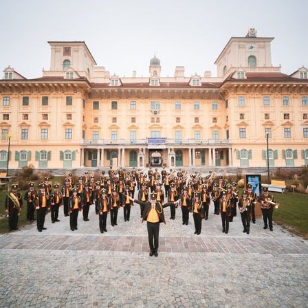 Eine Brassband steht vor einem großen, prächtigen Gebäude. Die Mitglieder tragen Uniformen und halten Instrumente. Die Umgebung ist formell und sonnig.