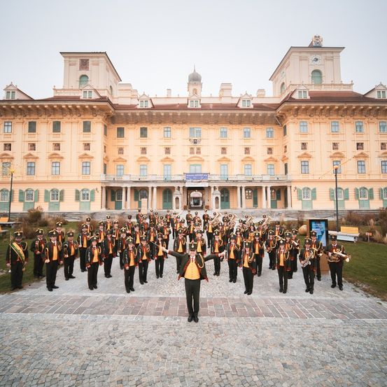 Eine Brassband steht vor einem großen, prächtigen Gebäude. Die Mitglieder tragen Uniformen und halten Instrumente. Die Umgebung ist formell und sonnig.