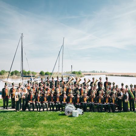 Ein Marschkapelle steht in Formation auf einem Rasen am Wasser, mit Booten und einer entfernten Landschaft im Hintergrund.