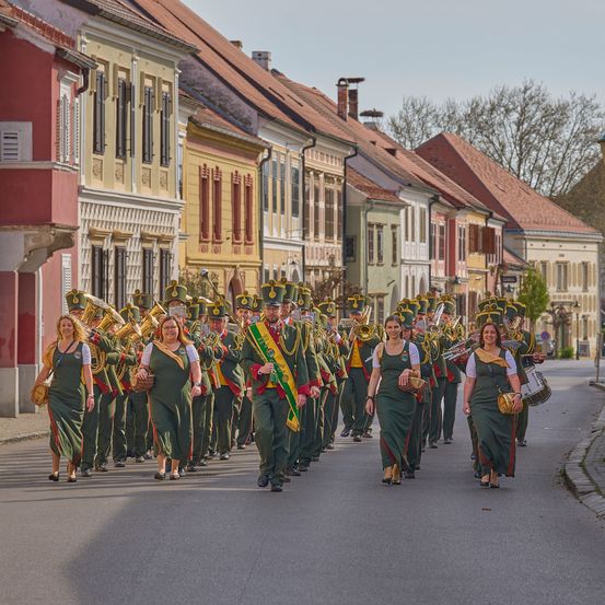 Eine Musikkapelle in grünen Uniformen geht in einer Stadt die Straße entlang. Bäume und Gebäude säumen die Straße.
