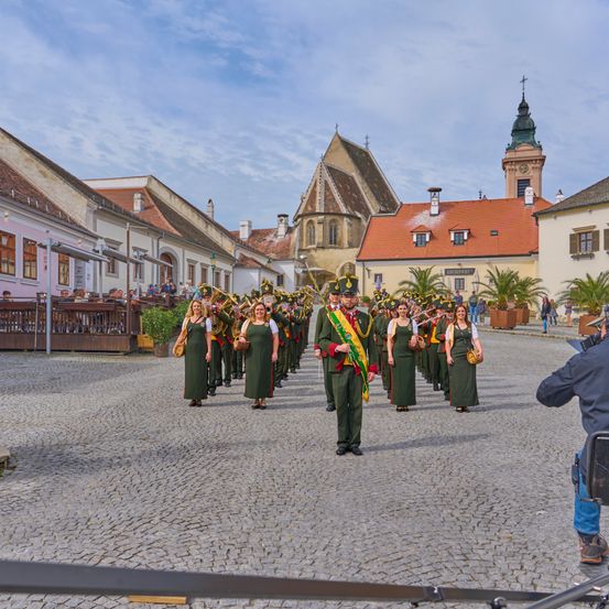 Eine Musikkapelle in grünen Uniformen tritt auf einer Kopfsteinpflasterstraße in einer historischen Stadt auf, mit Zuschauern und Gebäuden im Hintergrund.