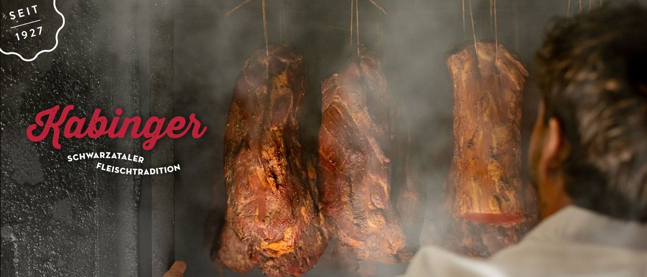 Mehrere Stücke gepökeltes Fleisch hängen an Schnüren in einer rauchigen Umgebung. Das Fleisch hat eine satte, rotbraune Farbe, was auf eine Räucherung hinweist.