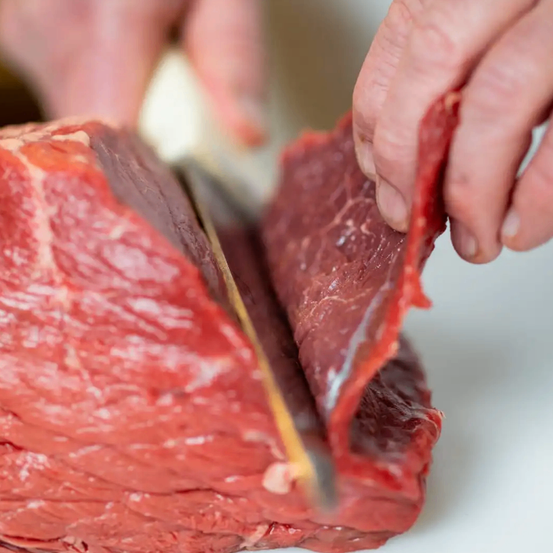 Eine Person schneidet ein großes Stück rotes Fleisch mit einem Messer auf einer weißen Oberfläche. Das Fleisch wird in dünne Scheiben geschnitten.