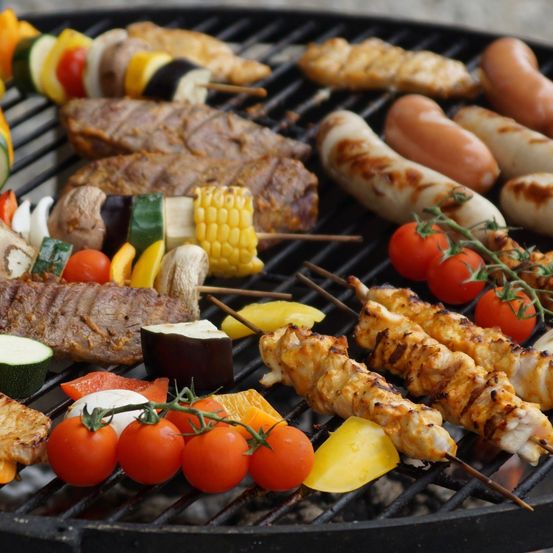 Ein Grill ist mit verschiedenen Fleischsorten, Gemüse und Würsten gefüllt. Hühnerspieße, Würste und Rindfleisch sind auf dem Grill angeordnet. Tomaten, Mais und verschiedenes Gemüse sind ebenfalls vorhanden.