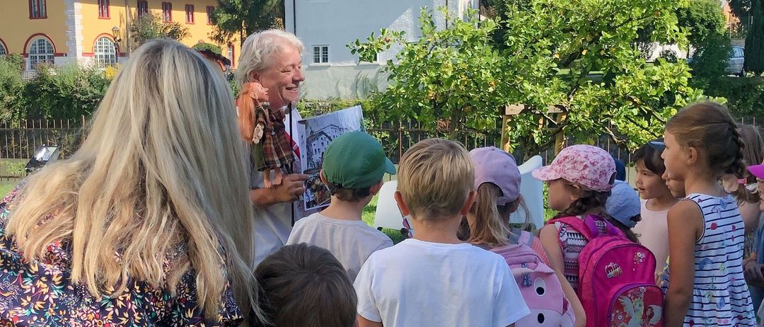 Eine Gruppe von Kindern und einer Frau hört einem Mann zu, der ein Buch und eine Puppe in der Hand hält. Sie sind draußen in der Nähe eines weißen Gebäudes und eines grünen Baumes.