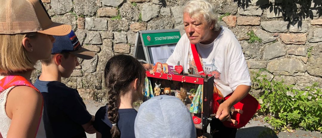 Eine ältere Frau zeigt einer Gruppe von Kindern ein kleines Puppentheater vor einer Steinmauer.
