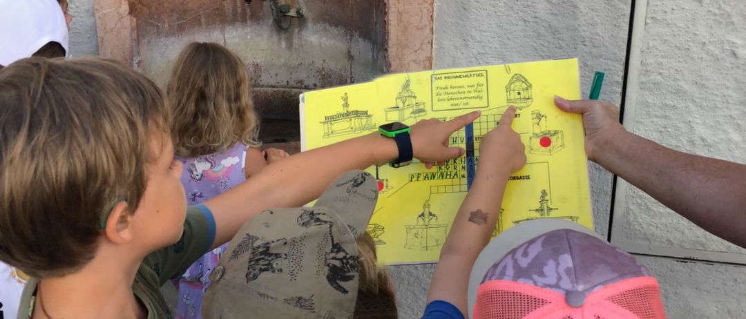 Kinder versammeln sich um einen Brunnen, ein Kind zeigt auf eine gelbe Karte, während es eine Mütze trägt. Die Karte hat Text und einen roten Punkt. Ein Mädchen in einem rosa Kleid schaut auf die Karte.