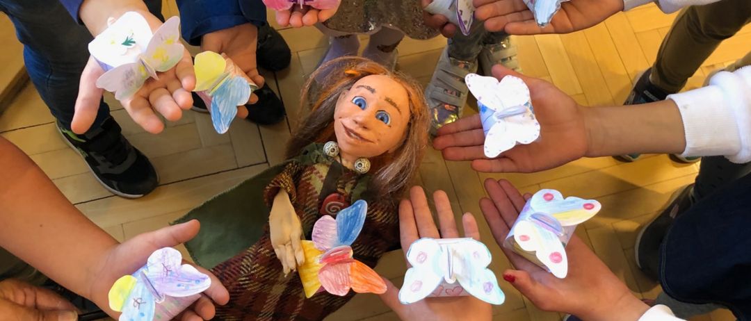 Eine Gruppe von Kindern ist um eine Puppe versammelt und hält bunte Schmetterlingsausschnitte in den Händen. Die Puppe ist in einem gemusterten Outfit gekleidet und hat gemalte Augen. Die Kinder tragen verschiedene Schuhe, und der Boden ist aus Holz.