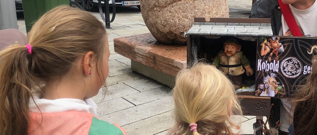 Eine ältere Frau zeigt einer Gruppe von Kindern in einer Stadt ein Puppenspiel. Die Aufführung findet auf einer kleinen Bühne statt. Hinter ihnen sind geparkte Autos und Gebäude.