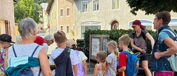 Eine Gruppe von Menschen, darunter Kinder, steht auf einer Kopfsteinpflasterstraße in einem Dorf. Sie tragen Rucksäcke und einige haben Hüte. Eine Frau schiebt einen Kinderwagen. Gebäude mit Fensterläden und Grünpflanzen sind im Hintergrund.
